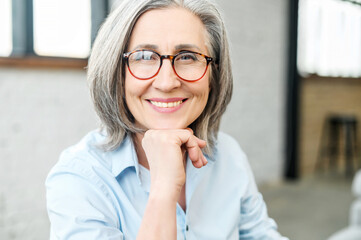 Headshot portrait of a charming beaming elegant mature woman with her hand on a chin, wearing glasses and looking at the camera. Close-up of a middle-aged gray-haired female office worker or a teacher