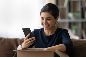 Fototapeta na wymiar Happy young Indian woman unbox parcel with internet order, track or rate good delivery service on smartphone. Smiling ethnic female buyer client shop online on cellphone. Shipping concept.