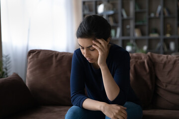 Fototapeta na wymiar Sad young Indian woman sit on sofa at home feel distressed depressed having life or relationship problems. Upset millennial mixed race female suffer from headache migraine. Depression concept.
