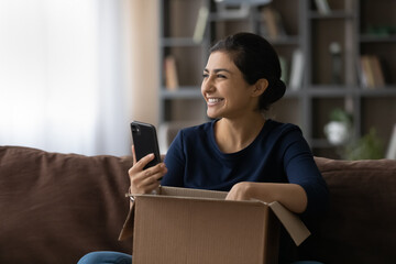 Overjoyed young Indian woman open unpack box with order shopping online on cellphone from home....
