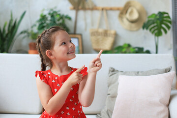 Cute little girl sitting on cozy sofa and pointing up on something intresting by fingers.