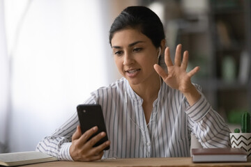 Young Indian woman in headphones use cellphone talk on video call with client or customer. Millennial mixed race female in earphones have webcam online conference on smartphone. Virtual event concept.