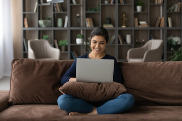 Happy millennial Indian woman sit relax on couch in living room study distant on computer gadget. Smiling young ethnic female rest on sofa at home office work online on laptop. Technology concept.