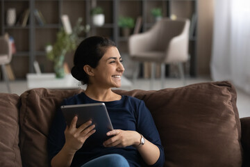 Smiling young Indian woman sit relax on couch in living room use tablet look in distance dreaming...