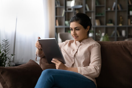 Millennial Indian Woman Sit Relax On Sofa In Living Room Look At Tablet Screen Talk On Video Call Online. Young Mixed Race Female Use Pad Gadget Browse Wireless Internet On Web. Technology Concept.