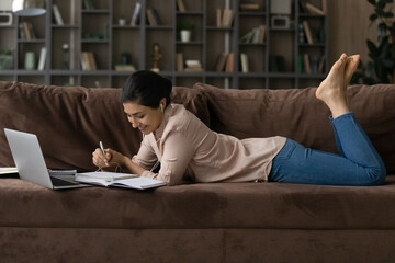 Happy millennial teenage Indian female lying on couch in living room study online on computer. Smiling mixed race woman watch webinar on laptop, take course or training on internet from home.