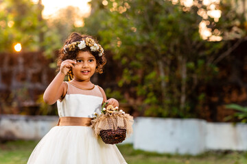 Portrait of an Asian, Indian female child dressed as a flower girl in a white dress and floral hair band and holding a bouquet outdoors in a flower park