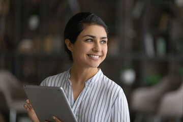Smiling Indian woman use modern tablet gadget look in distance planning thinking. Happy millennial ethnic female user or customer browse wireless internet on pad. Technology, communication concept.