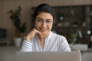 Fototapeta premium Close up of smiling young Indian woman in glasses look at computer screen work online at home office. Happy millennial mixed race female use laptop talk on video call with client. Technology concept.