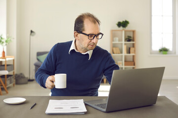 Balding middle aged man using laptop. Freelance worker or online tutor sitting at desk in home office, drinking coffee and working on computer. People browsing Internet, e-learning, remote job concept