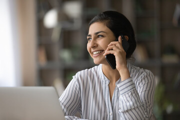 Smiling young Indian woman work on laptop talk speak on modern smartphone. Happy millennial mixed...