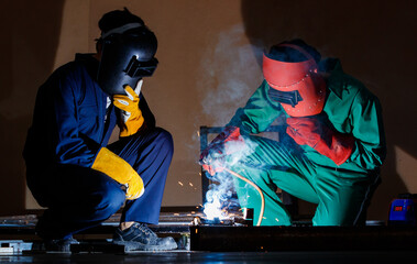 two engineers working in the dark at nighttime. Mechanics wearing a mechanic coveralls work together to weld the metal rod, making nice spark, bokeh, and smoke