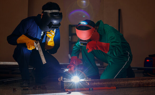 two engineers mechanics sitting and working in a workshop of a factory. They are helping each other to weld a piece of metal rod with a welding machine at night time