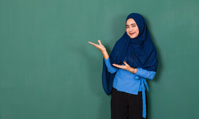 a pretty confident Asian Muslim woman dressed nicely wearing blue hijab posing for photographing acting introducing something in a copy space over green background