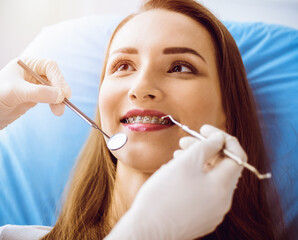 Smiling young woman with orthodontic brackets examined by dentist in sunny dental clinic. Healthy teeth and medicine concept