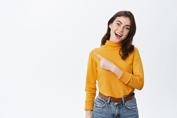 Hey check this out. Cheerful caucasian woman in yellow sweater pointing finger left. Girl showing advertisement with happy smiling face, standing on white background