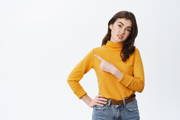 Skeptical and unamused young woman look at camera doubtful, pointing finger left at average, bad promo offer, standing against white background