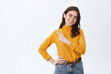 Fototapeta premium Young beautiful woman pointing finger left at promo, smiling and staring at camera, showing advertisement, demonstrating copy space, white background
