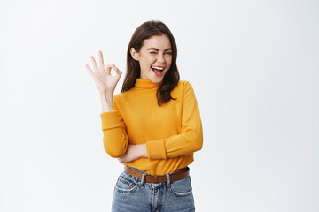 Confident young woman winking to assure you, showing okay sign and smiling, guarantee good quality, say alright or agree, like something or recommending, white background