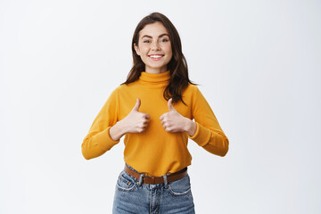 Young excited woman showing full support, thumbs up and smiling in approval, say yes, agree or approve. Girl likes good choice, recommending product, white background