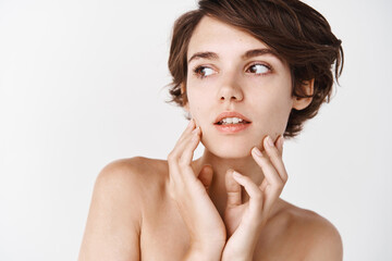 Close-up of tender young woman with naked shoulders, gently toughing natural no makeup facial skin, looking away pensive, standing over white background