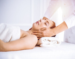 Obraz na płótnie Canvas Beautiful brunette woman enjoying facial massage with closed eyes in sunny spa center . Relaxing treatment and cosmetic medicine concepts