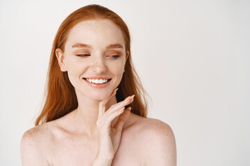 Skincare. Close-up of redhead woman with pale soft skin, smiling white teeth and touching clean no makeup face, looking aside, standing over white background naked