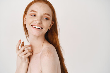 Close-up of pale redhead woman standing naked on white background, turn right and smiling at copy space about skincare - obrazy, fototapety, plakaty