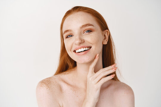 Beauty. Close-up of young beautiful redhead woman smiling at camera, touching perfect clean skin on face and looking happy, standing naked over white background