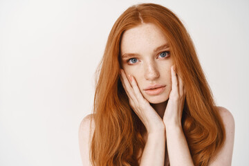 Beauty and skincare. Close-up of redhead pale woman touching face, looking at camera sensual, showing perfect no makeup skin, standing over white background