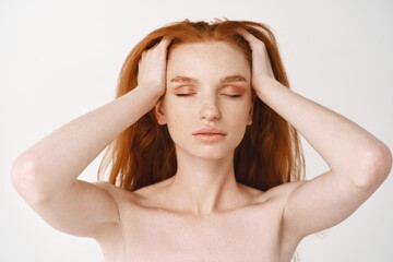Naklejka premium Close-up of relaxed young redhead woman with pale skin and freckles, massaging natural red hair with closed eyes, standing naked without make-up on white background