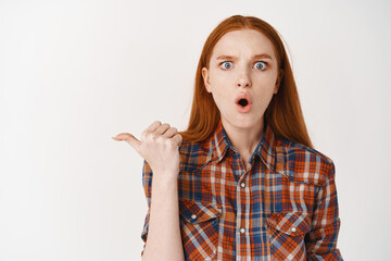 Shocked redhead woman with pale skin and no makeup, pointing finger left at banner, gasping amazed at camera, standing over white background