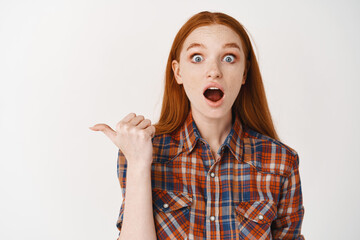 Amazed young female student checking out an advertisement, looking at camera with opened mouth, pointing finger left at promo banner, white background
