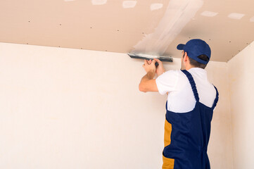 A man uses a spatula to putty a drywall joint on the ceiling