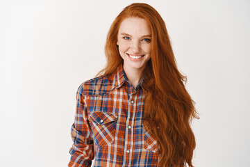 Obraz premium Image of attractive female model with long healthy red hair, smiling at camera confident. Young redhead woman standing in plaid shirt against white background