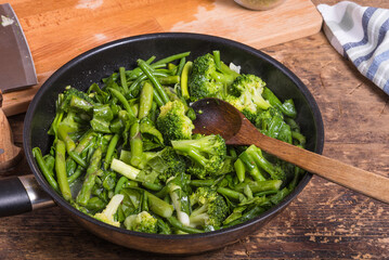 Cooking green vegetables for pasta in a frying pan - beans, broccoli, asparagus in a frying pan on a wooden table, tomatoes and chopped spinach on a board