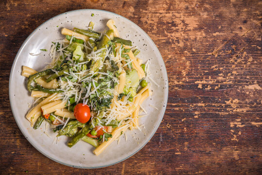 Vegetarian american and italian primavera pasta dish with broccoli, beans, asparagus, peas and tomatoes in a plate on a wooden table, top view, copy space