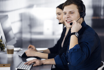 Two young people in headsets are talking to the clients, while sitting at the desk in a modern office. Focus on man in a blue shirt. Call center operators at work