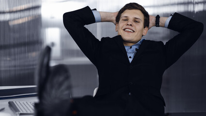 Friendly businessman in a black suit is sitting at the desk at his workplace. Portrait of a young specialist in a modern office. Concept of a success in a business