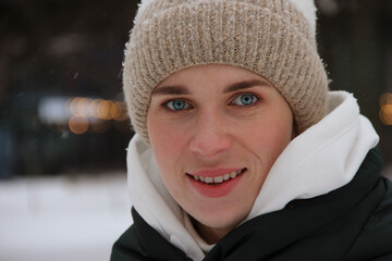 Close-up of the face of a smiling blue-eyed girl in a winter hat against the background of a winter park and lights