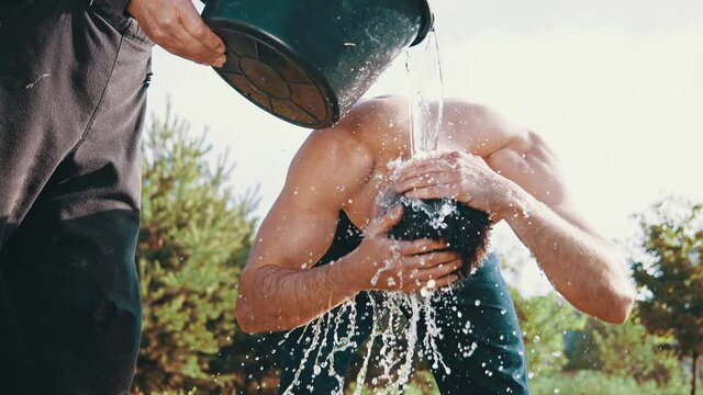 Man freshen up muscular caucasian guy with cool water out of the wooden bucket. Cool down, healthy lifestyle, strongman concept. Man freshing at street with green grass and trees at background