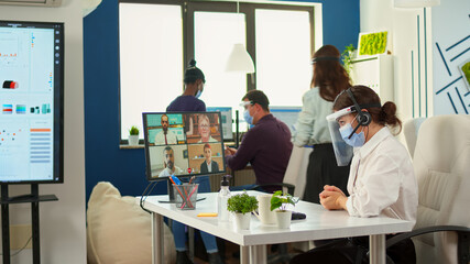 Manager with protection mask and visor having videomeeting discussing online using headset with remotely colleagues in business office. Multiethnic coworkers respecting social distance in company
