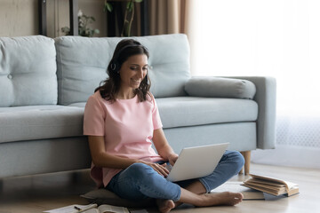 Obraz premium Concentrated indian lady sit on pillow on warm heated floor work on pc from home type report do research. Smiling arabian woman student in wireless headset study literature engaged in paperwork online