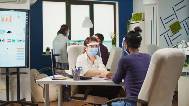 African employee and coworker with face masks planning financial project typing on computer and discussing sitting in business office with new normal. Multiethnic team respecting social distance