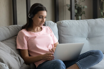 Confident arabic female in headset work from home online sit on sofa look at pc screen give client...