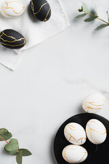 Frame of white and black Easter eggs decorated gold and eucalyptus leaves on marble table. Flat...
