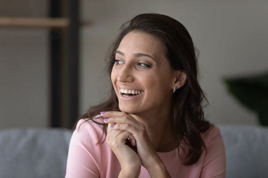 Head shot picture of happy positive indian arabic woman sitting on sofa laughing looking aside. Carefree young mixed race female relax at home laughing on funny joke demonstrating white healthy smile