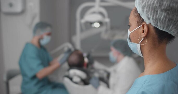 African woman intern standing in dental office watching professional dentist treating patient teeth