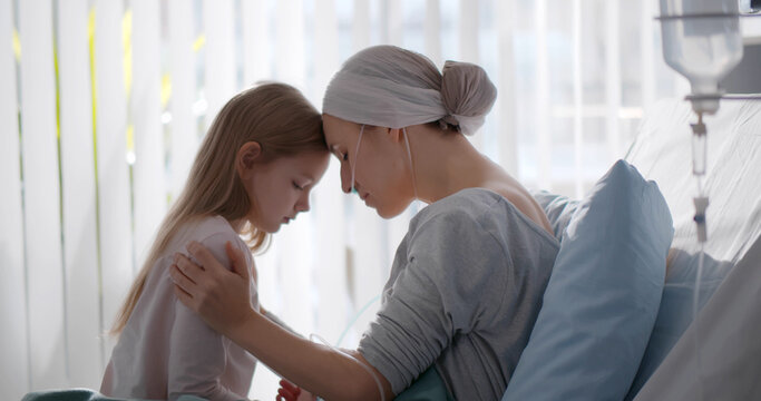 Side view of young woman with cancer sitting on hospital bed with little daughter
