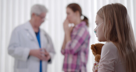 Fototapeta premium Sad sick kid with teddy bear sitting in hospital bed with doctor talking to upset mom on background